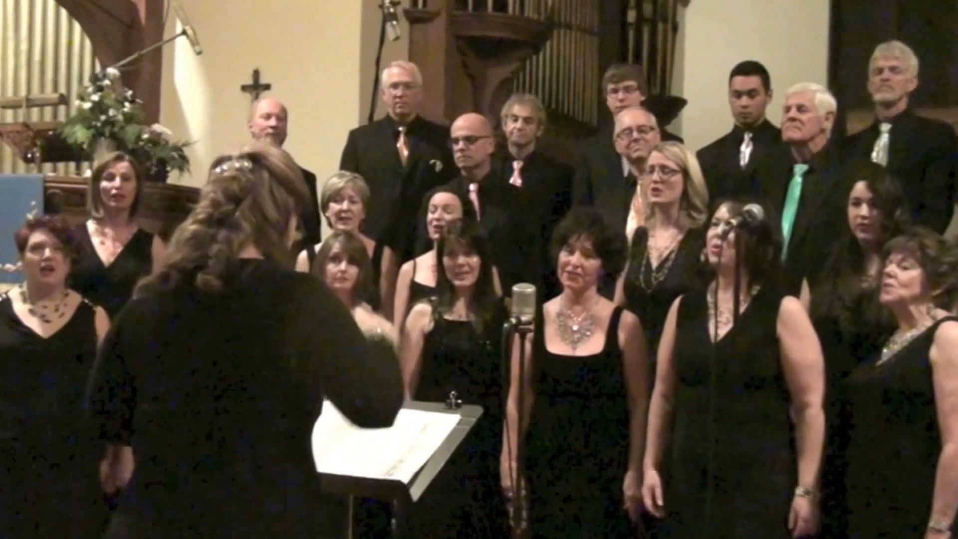 The choir in black, with coloured ties for the men, singing before the organ pipes and a Christmas flower arrangement, the conductor in the foreground with her back to the camera.