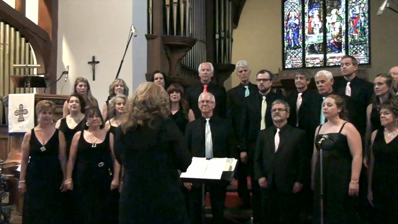 The choir in black concert dress beneath a stained-glass window, with Kim Kleineberg conducting.