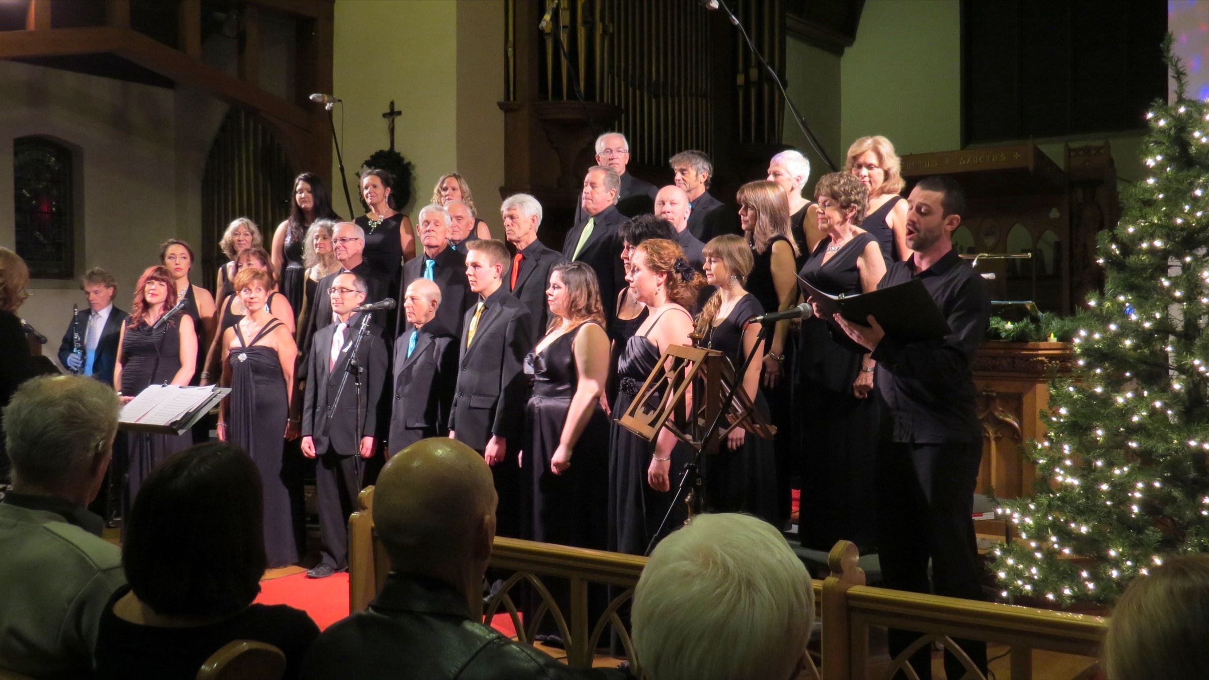 The choir in black on the chancel, the men in coloured ties, beside a lit Christmas tree; a male soloist at the front sings from a black folder, with the audience in the foreground.