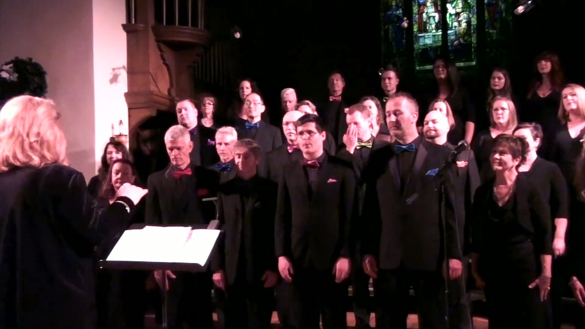 The choir in black concert dress with coloured bow ties, beneath a stained-glass window, with Kim Kleineberg conducting at the left.