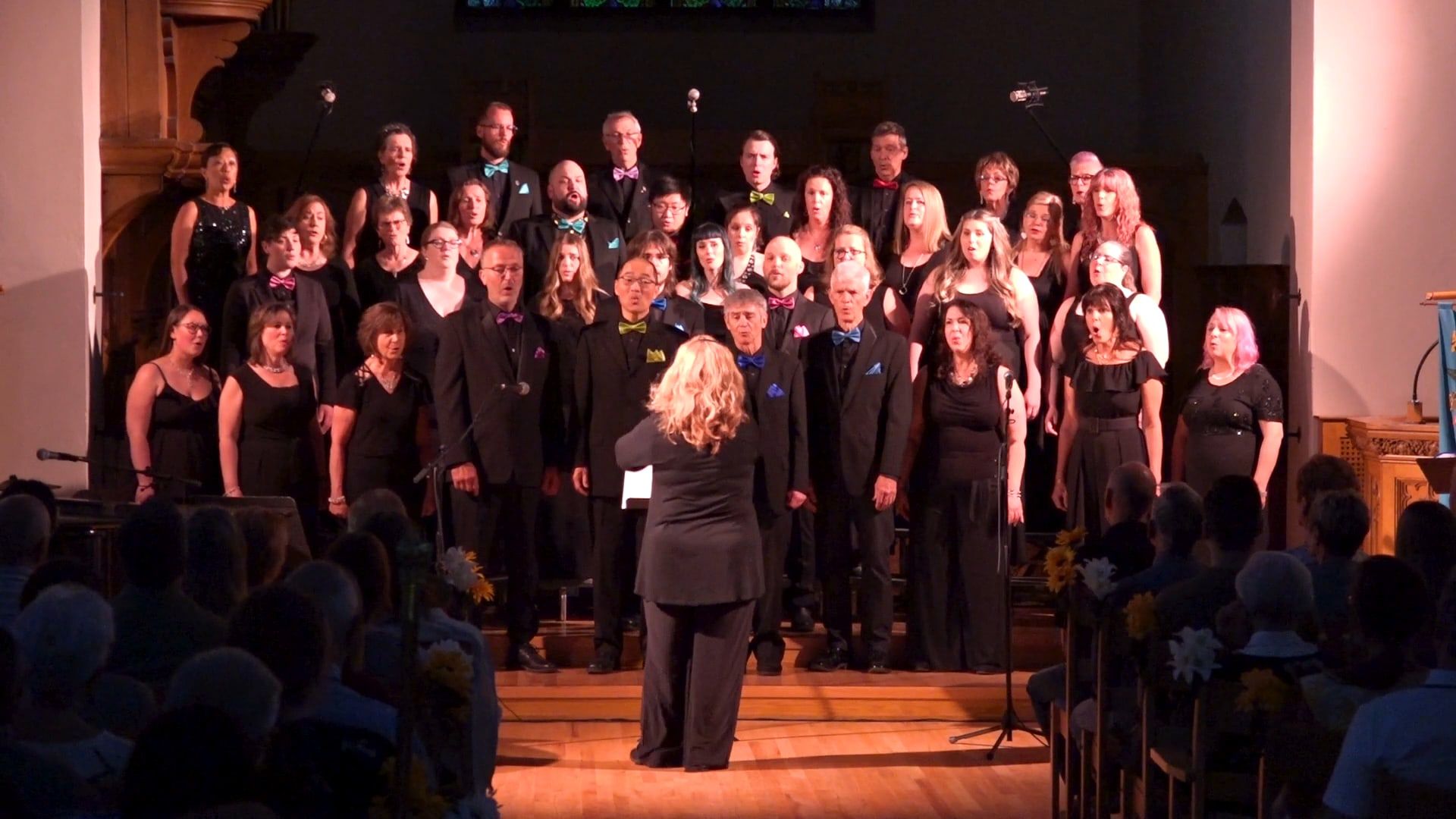 The choir on the risers in black, the men in bright bow ties, the conductor in the foreground and the audience in silhouette.