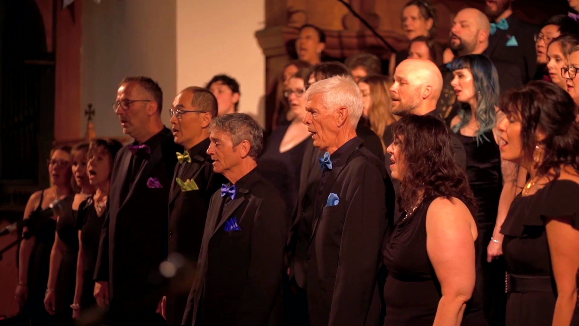 Rows of Spectrum Singers in black concert dress, each with a brightly coloured bow tie or pocket square, singing together in St Michael's Cathedral.