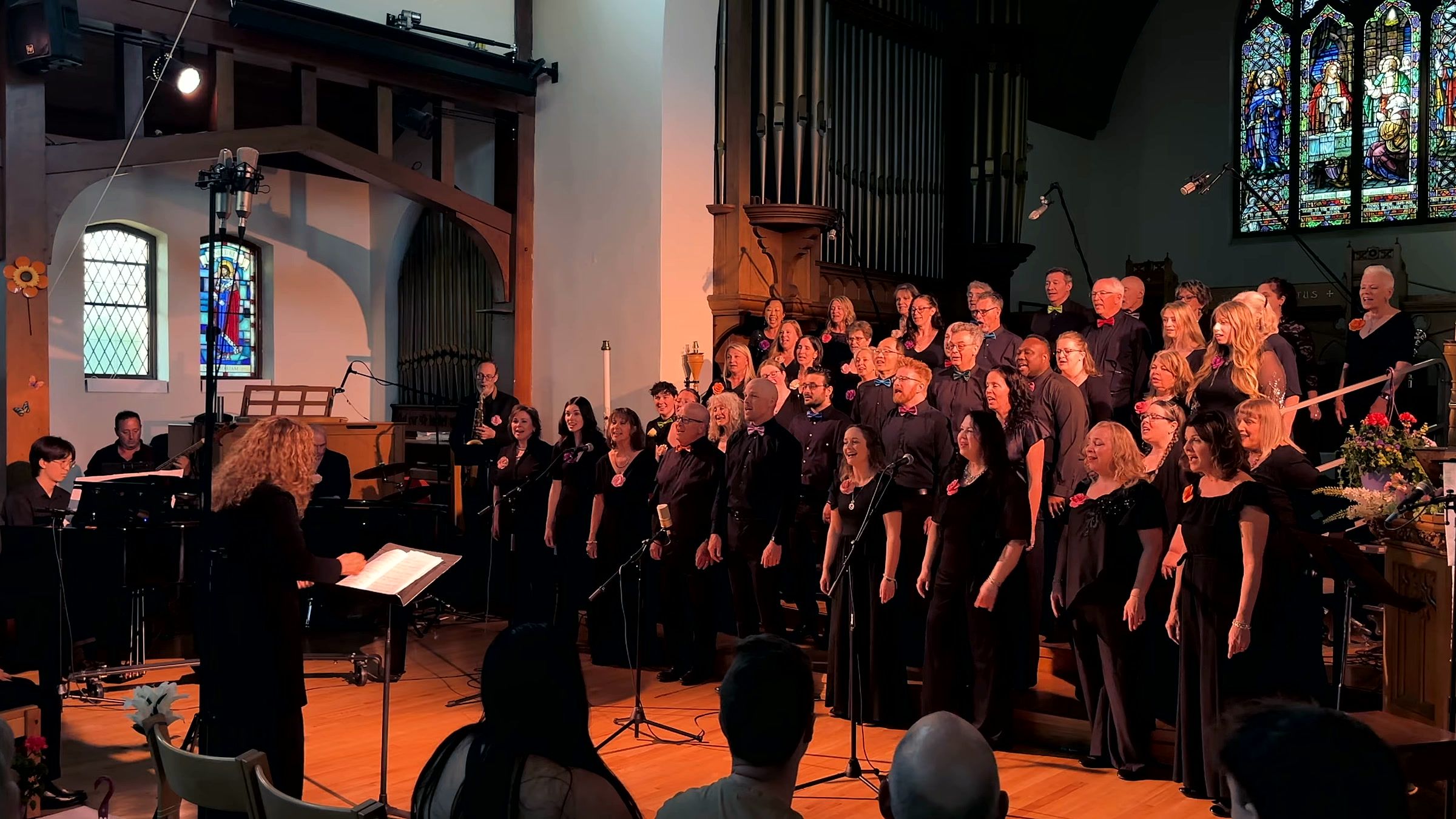 The Spectrum Singers in black concert dress singing on the steps beneath a stained-glass window, with Kim Kleineberg conducting at the left and the band behind her.