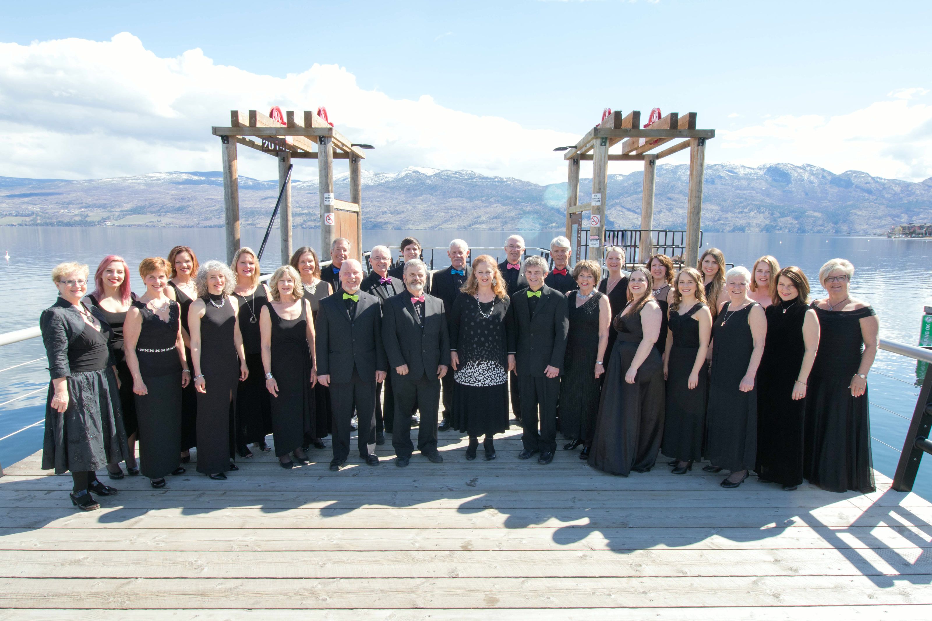 The full choir in black concert dress, posed on a wooden dock with a lake and snow-capped hills behind them on a sunny day.