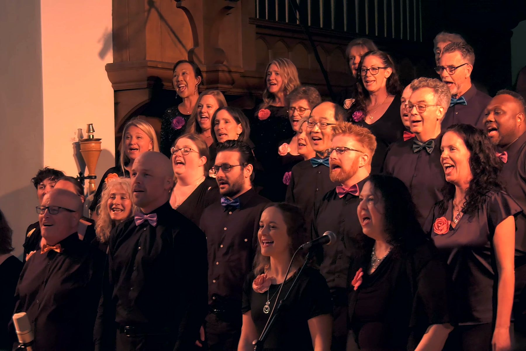 Rows of singers in black concert dress with coloured bow ties and fabric roses, singing and smiling mid-performance.
