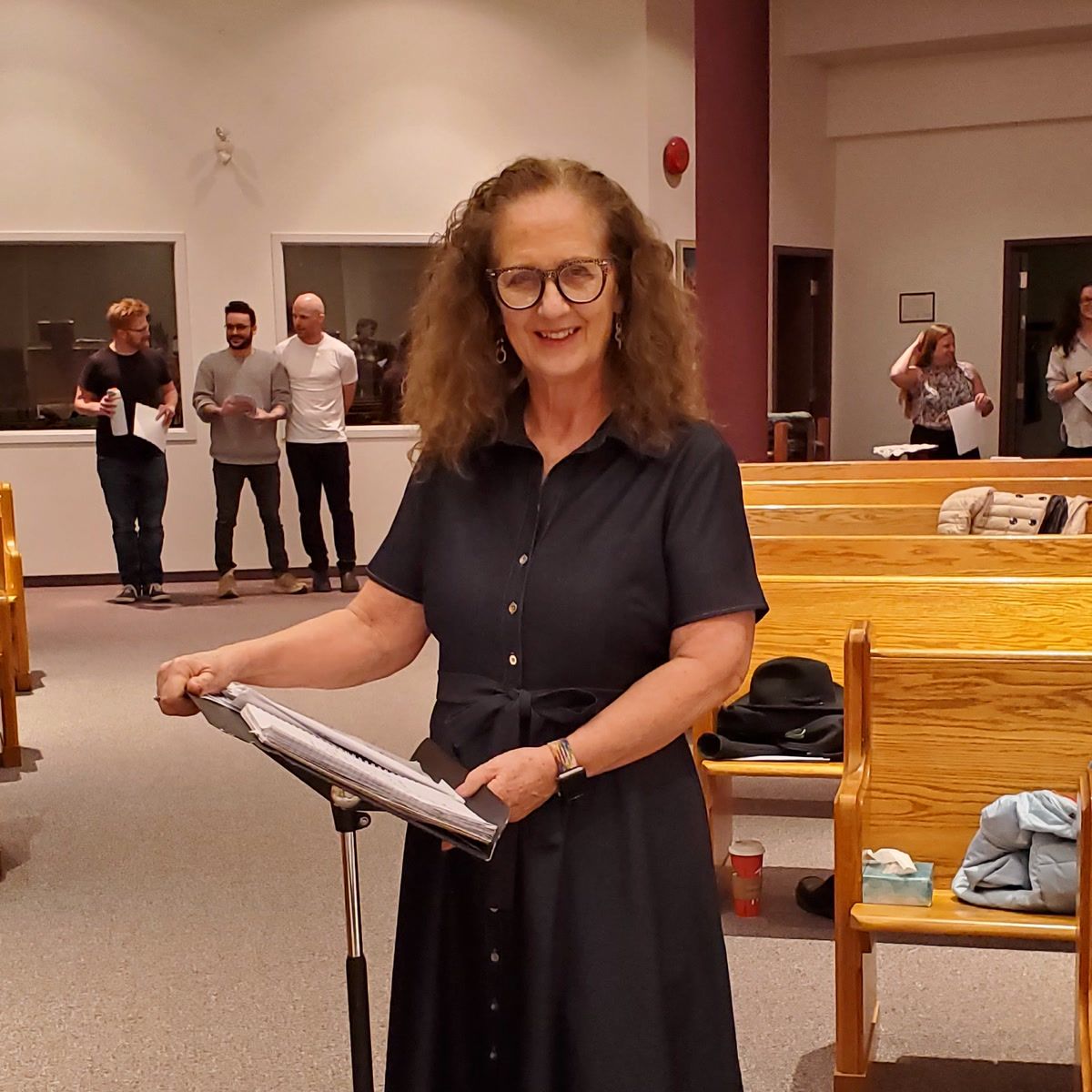 Kim Kleineberg, smiling, at a music stand among wooden pews, with singers holding sheet music behind her.